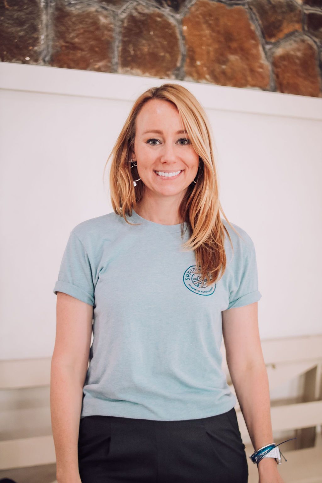 Woman smiling, wearing a blue t-shirt and black pants. Standing indoors, with a white wall backdrop.