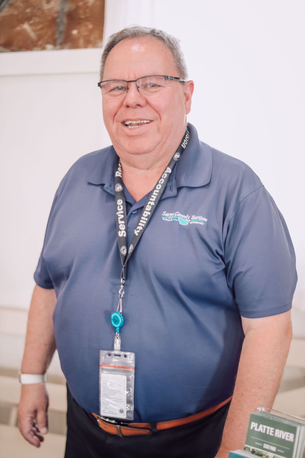 Man in blue polo shirt with lanyard smiling. Indoors, next to a white wall.