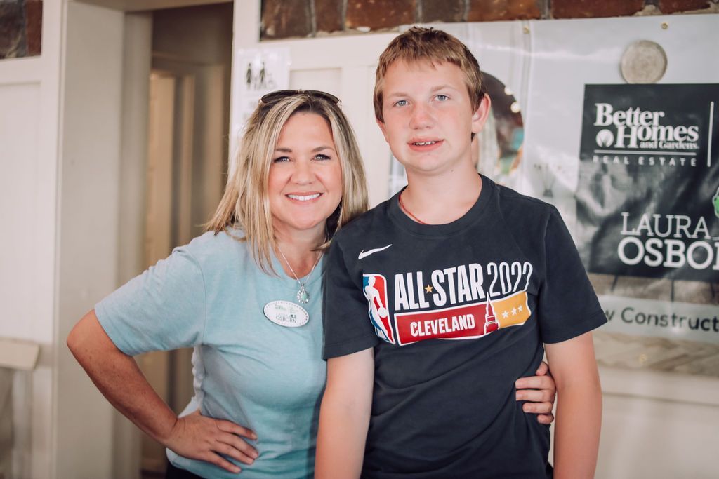 Woman and teen boy pose. Woman in light blue shirt smiles, hand on hip. Boy in black t-shirt smiles, arm around her.