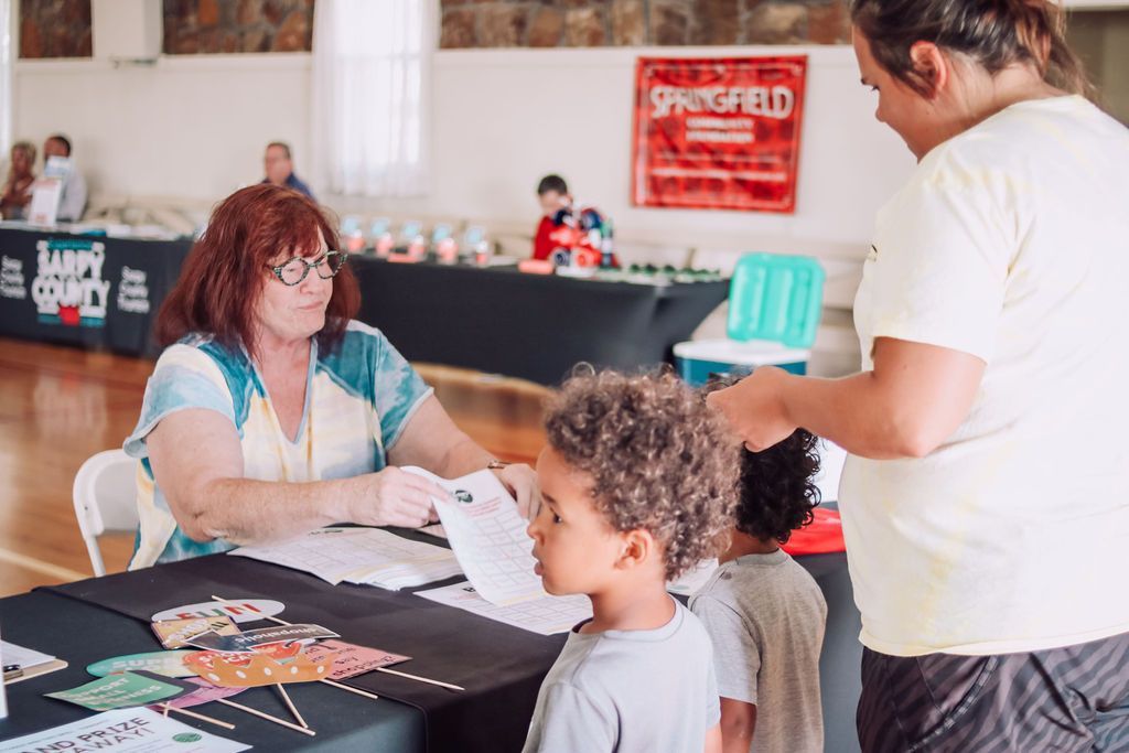 Woman at a table helping a child; another person stands nearby. Event in a room.