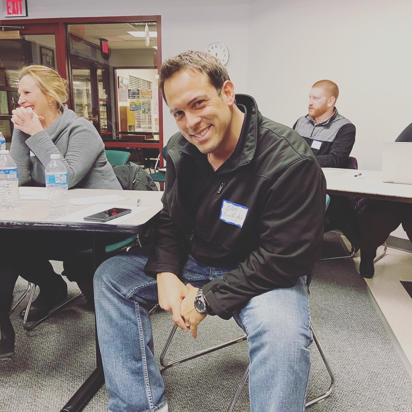 Man in black jacket and jeans smiling at the camera, seated at a desk in a classroom setting.