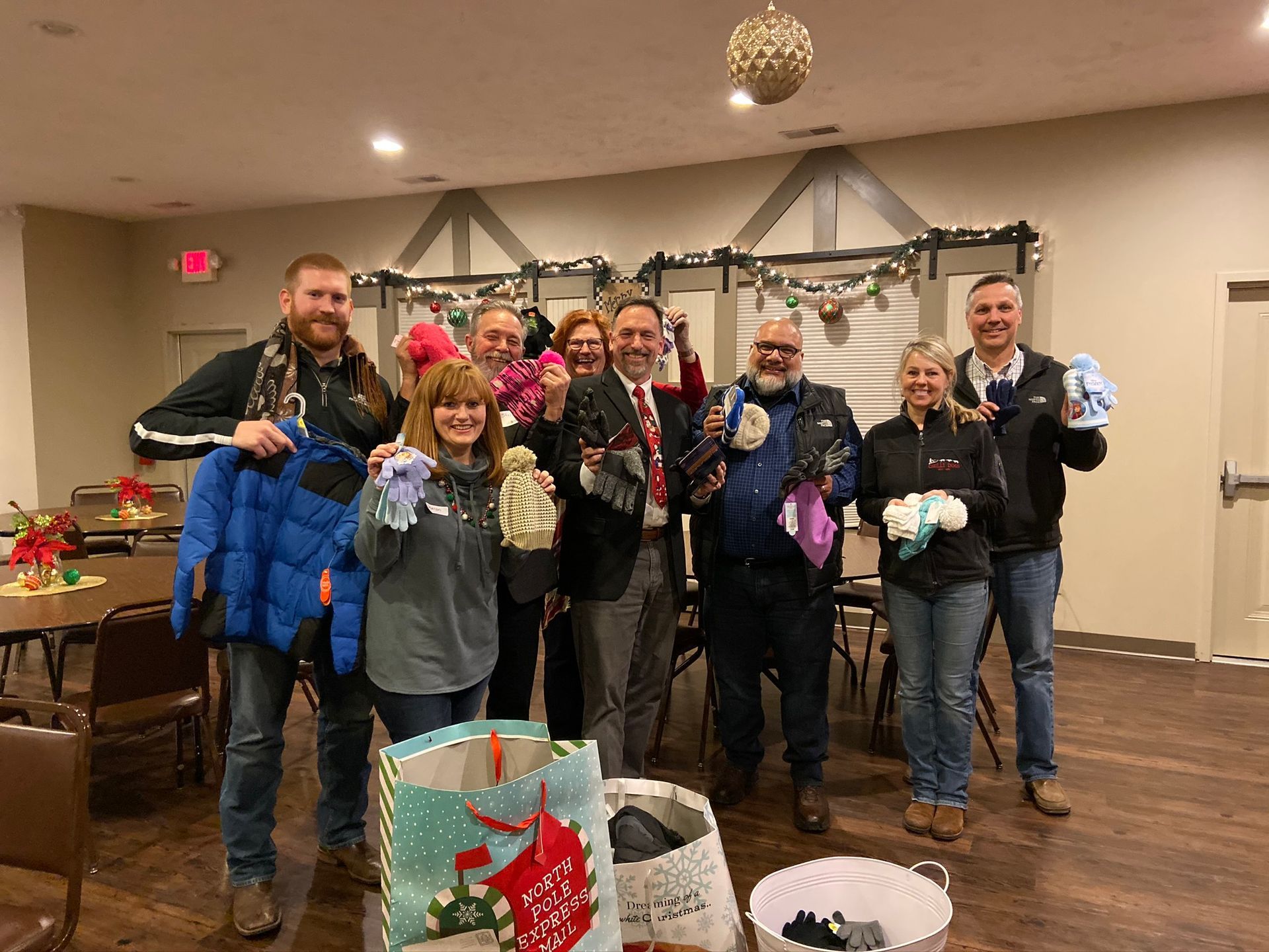 Group of people holding winter clothing, smiling in a decorated room.