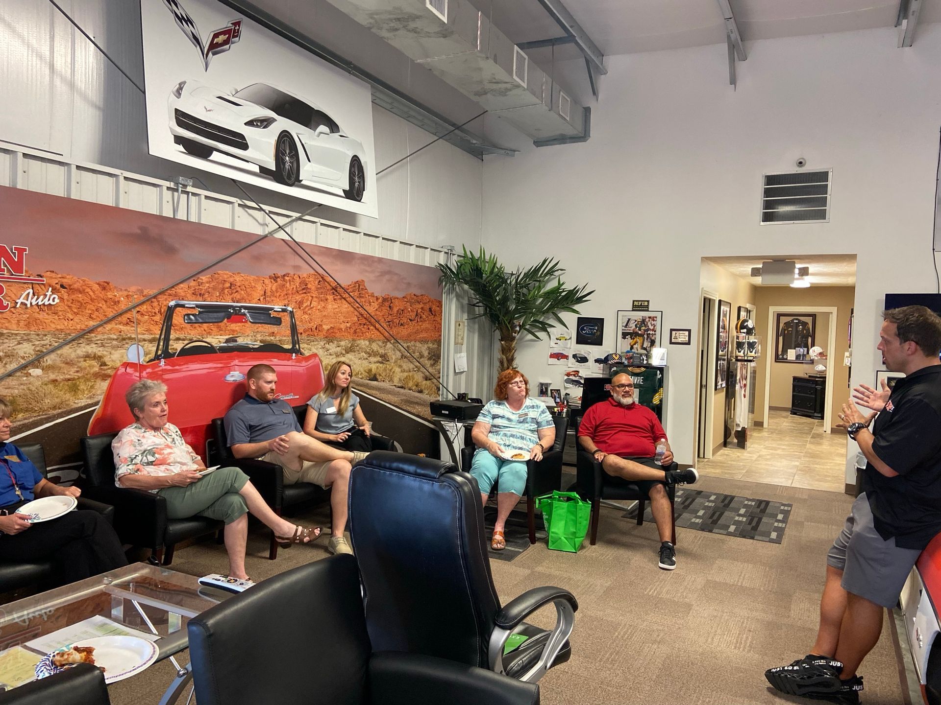 People seated in a room, listening to a man speak. Corvette decor visible.