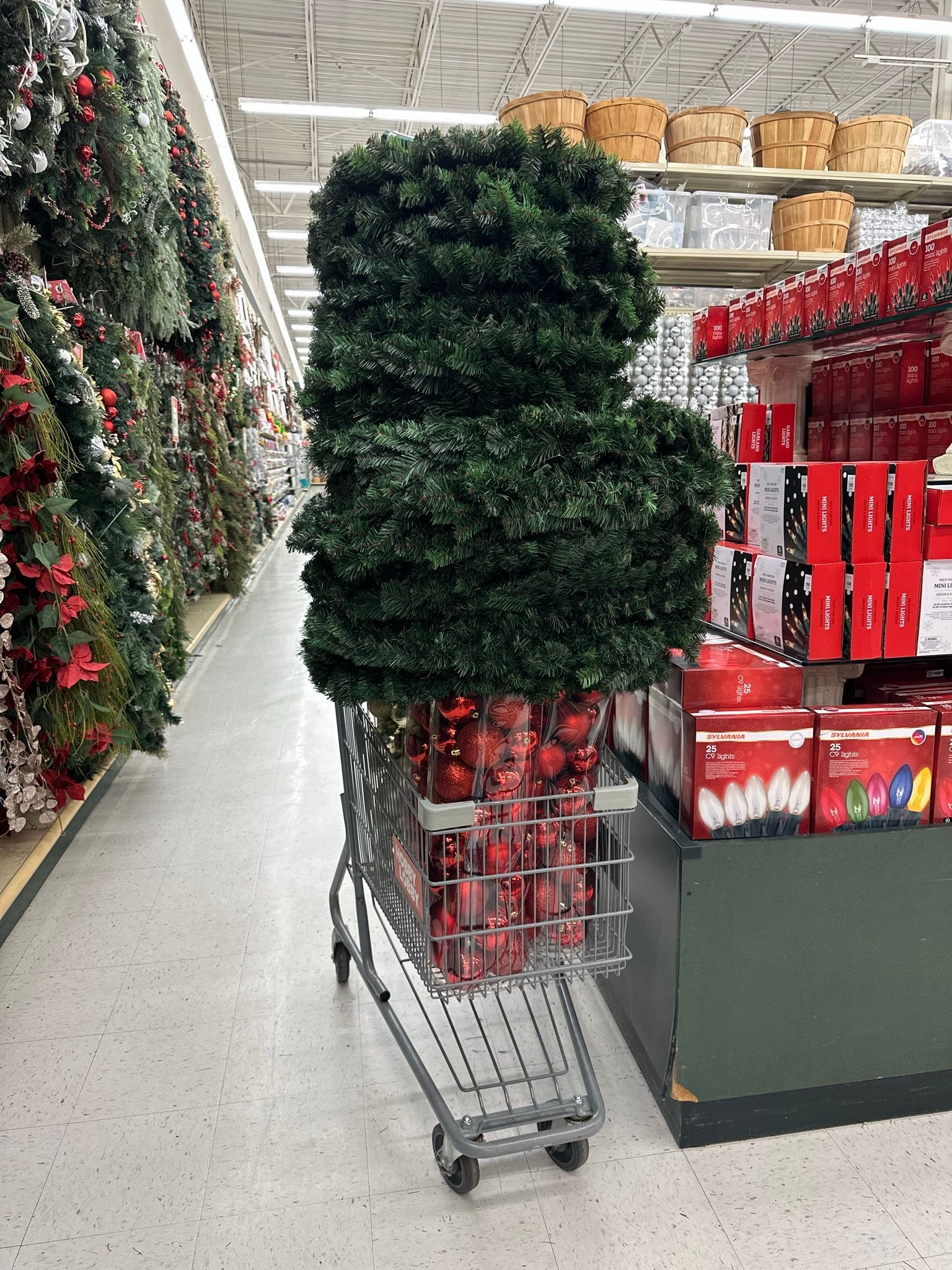 Shopping cart in store aisle filled with Christmas decorations, including green foliage and red ornaments.