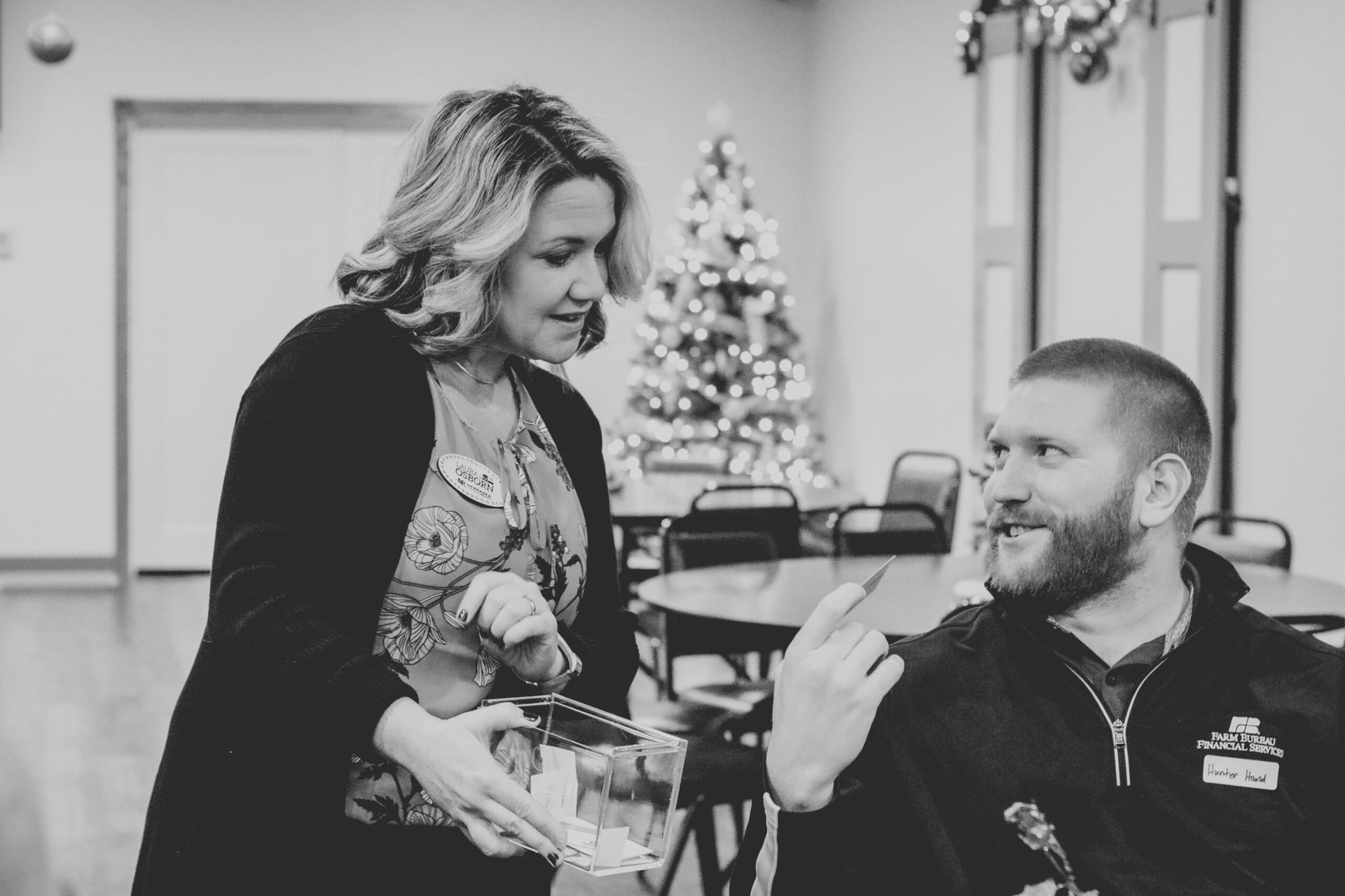 Woman offers item to man indoors with Christmas tree.