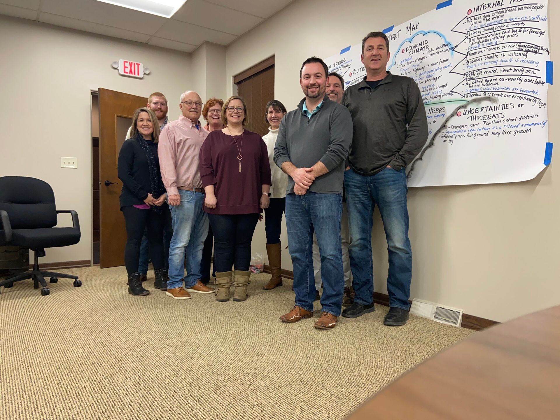 Group of people in an office. A whiteboard with writing is behind them. They are smiling at the camera.