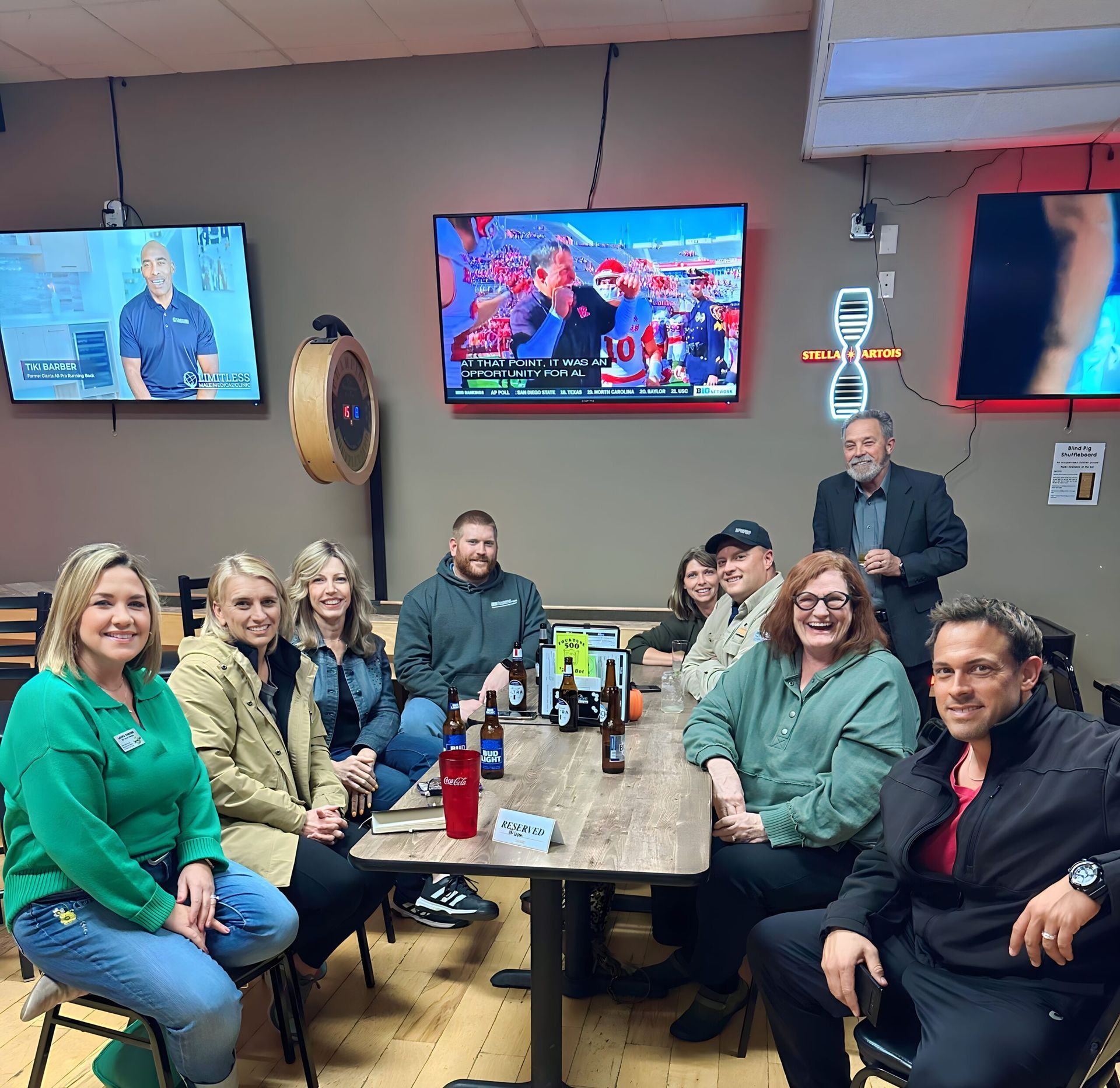 Group of people at a bar, seated around a table, watching TV screens.