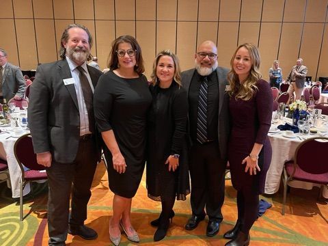 Five people pose for a photo in a banquet hall. They are dressed up and smiling. Tables are set in the background.