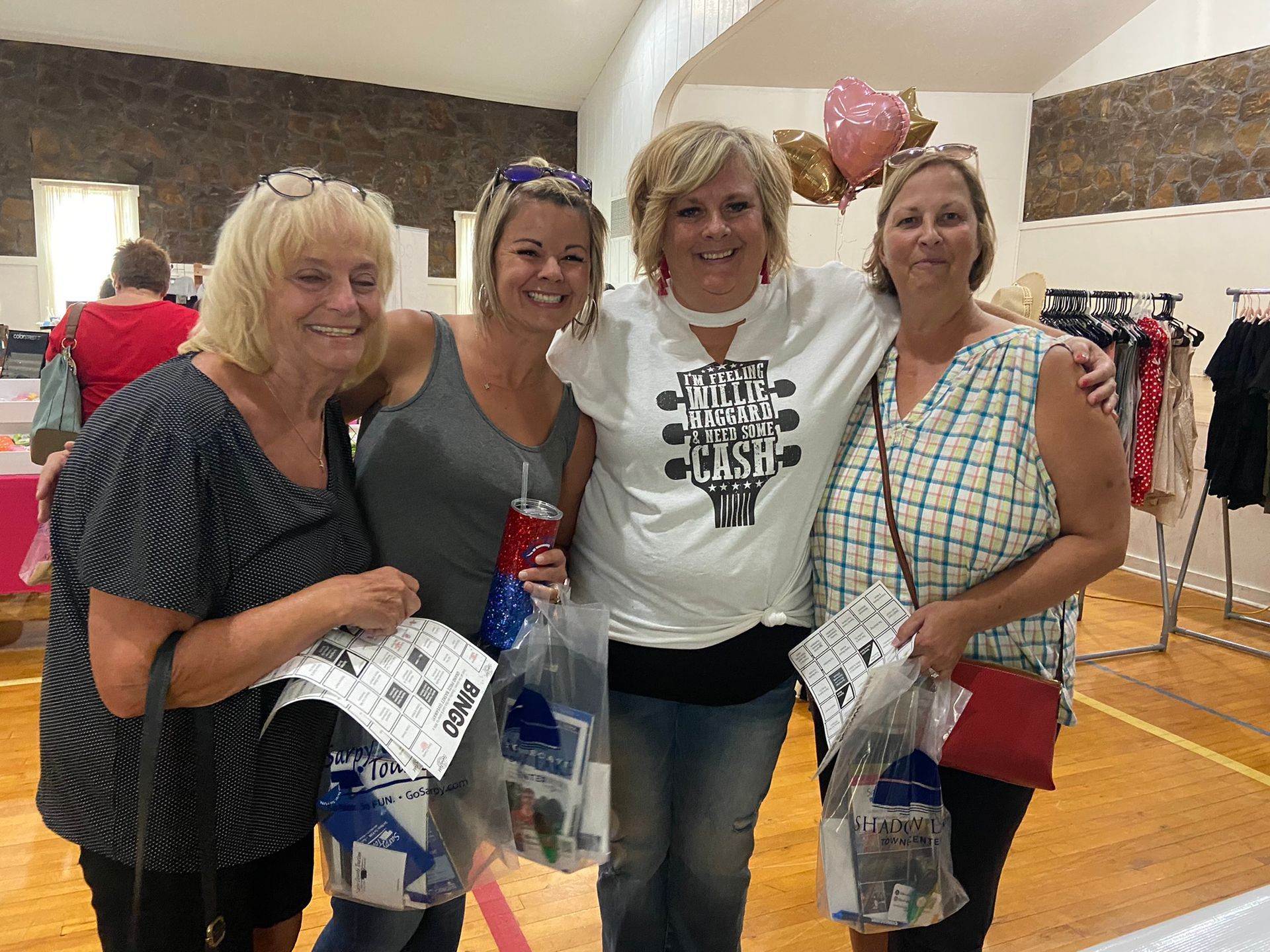 Four women smiling, holding bingo prizes. Inside a community hall with clothing racks and balloons.