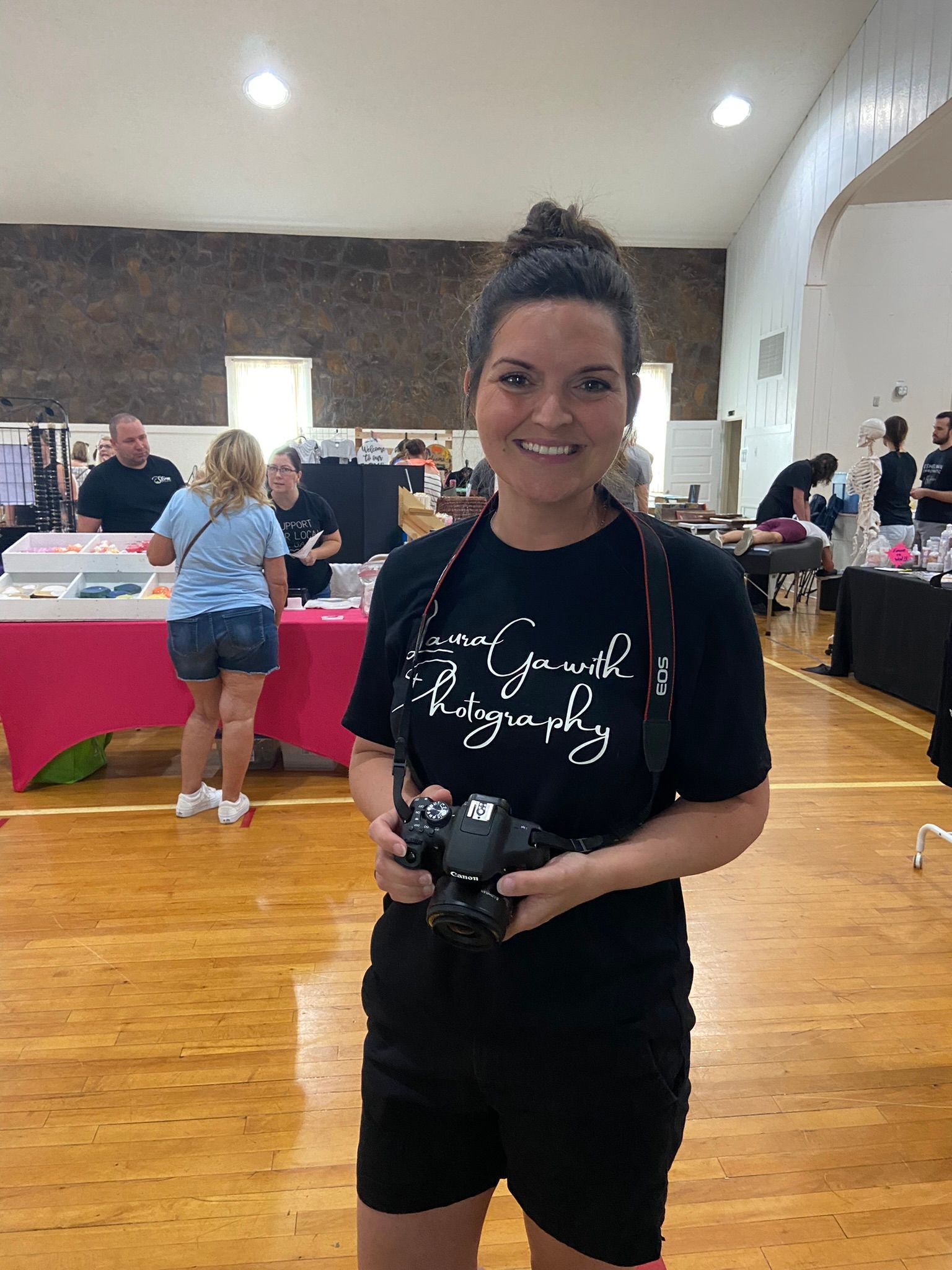 Woman with a camera at an event, wearing black, smiling. Event tables and people in the background.