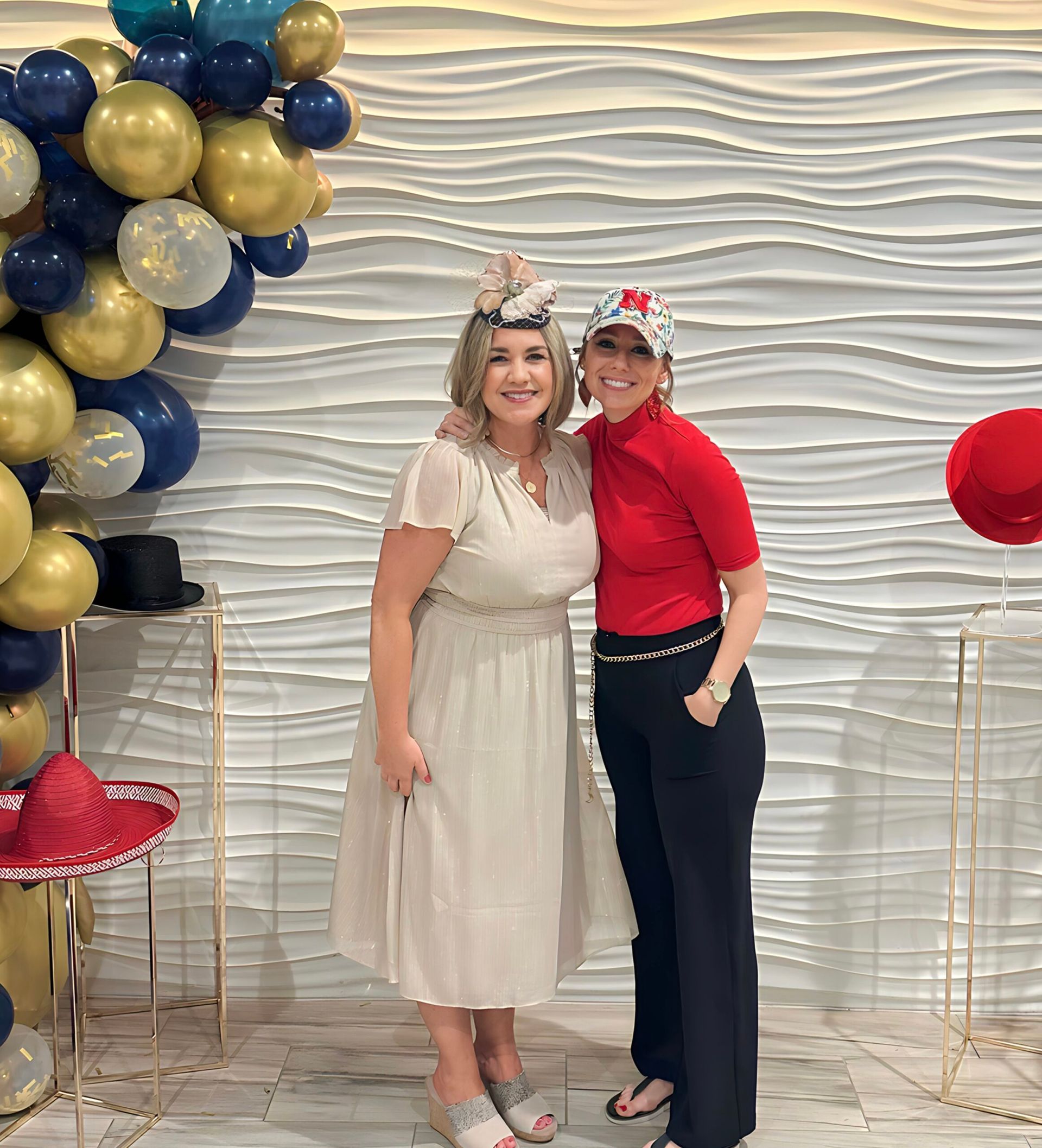 Two women smiling, posing for a photo. Balloons and hats decorate the backdrop.