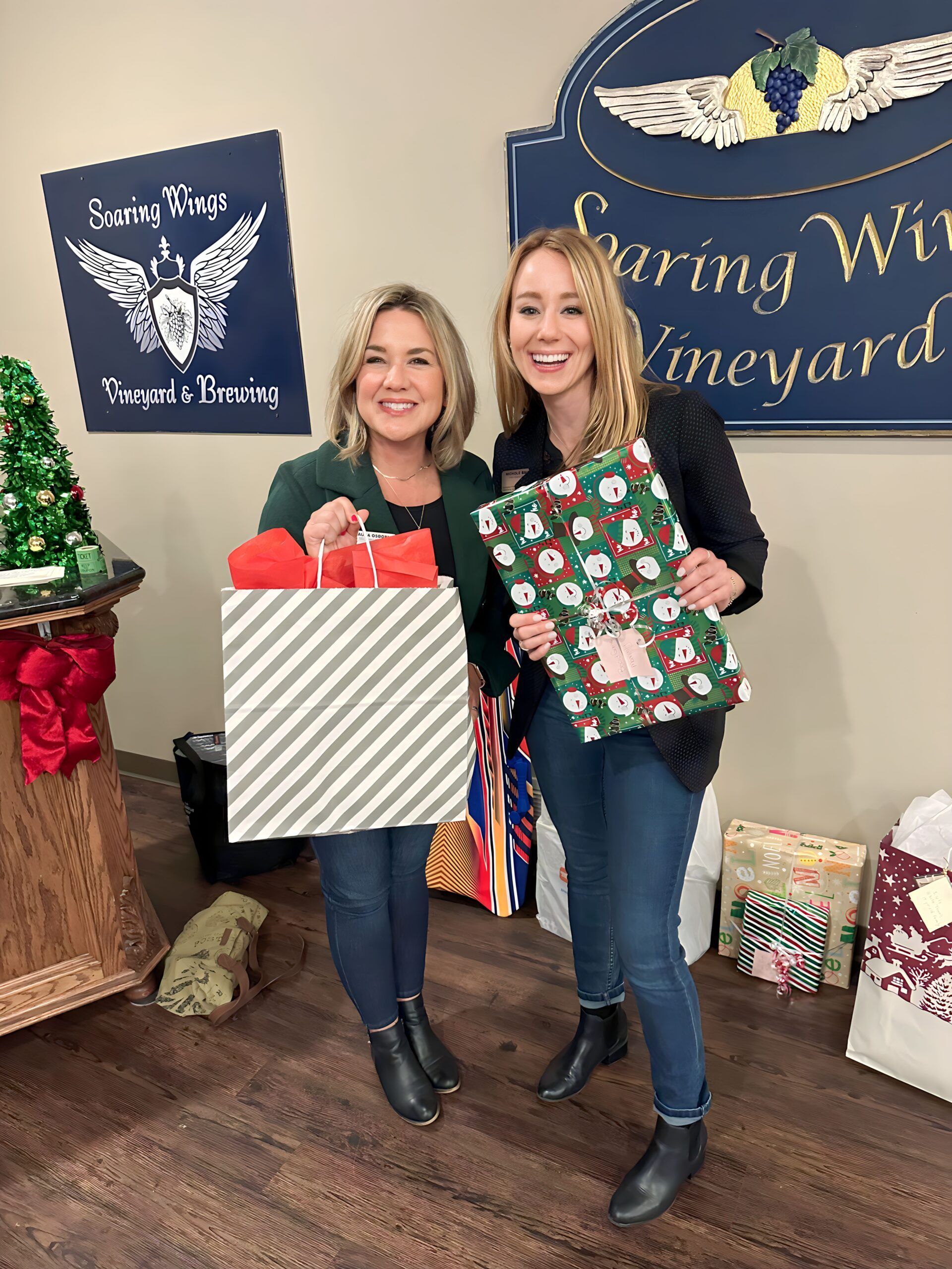 Two women smiling, holding wrapped gifts at a winery. Gray and green gift bags with holiday wrapping.