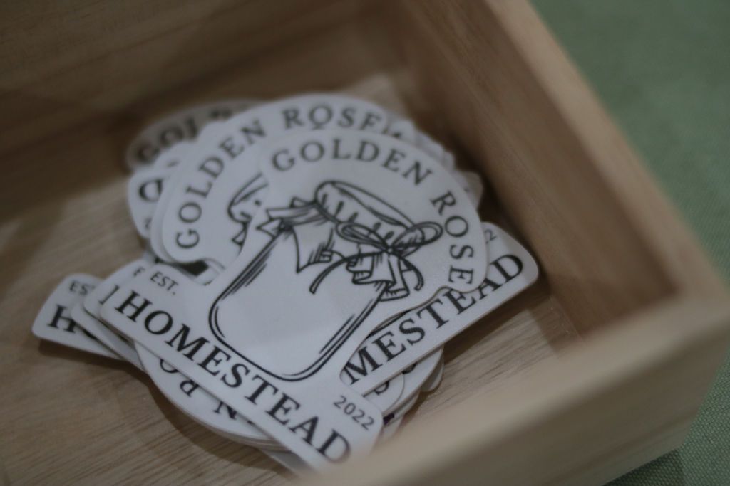 Pile of Golden Rose Homestead stickers in a wooden box, featuring a jar illustration and text.