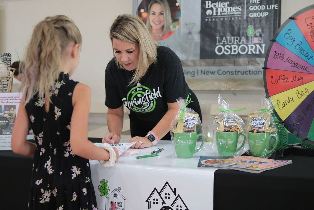 A woman signs something for a girl at a Better Homes and Gardens booth. Green mugs and prizes are on the table.
