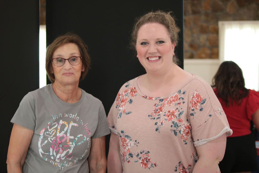 Two women smiling, standing near a black backdrop. One in a floral shirt, the other in a gray t-shirt.