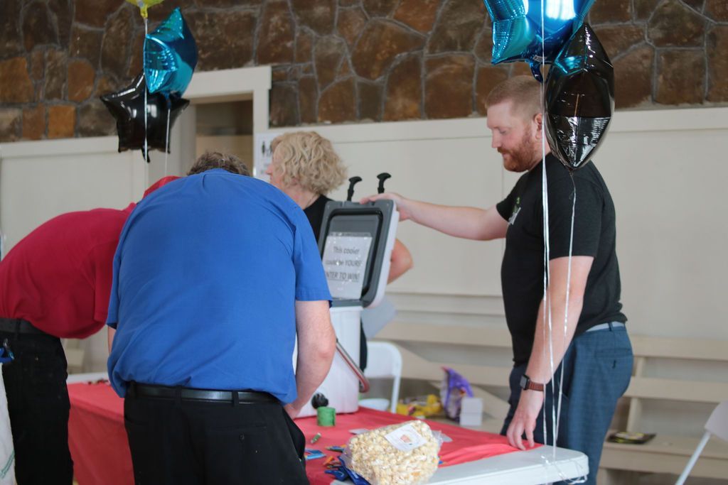 People at a table with a white machine, balloons overhead. A man in black touches the machine.