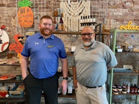Two men standing inside a shop; one in blue shirt, other in tan shirt. Shelves with goods visible.