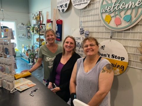 Three women standing behind a counter in a store. Smiling, surrounded by wall decor and merchandise.