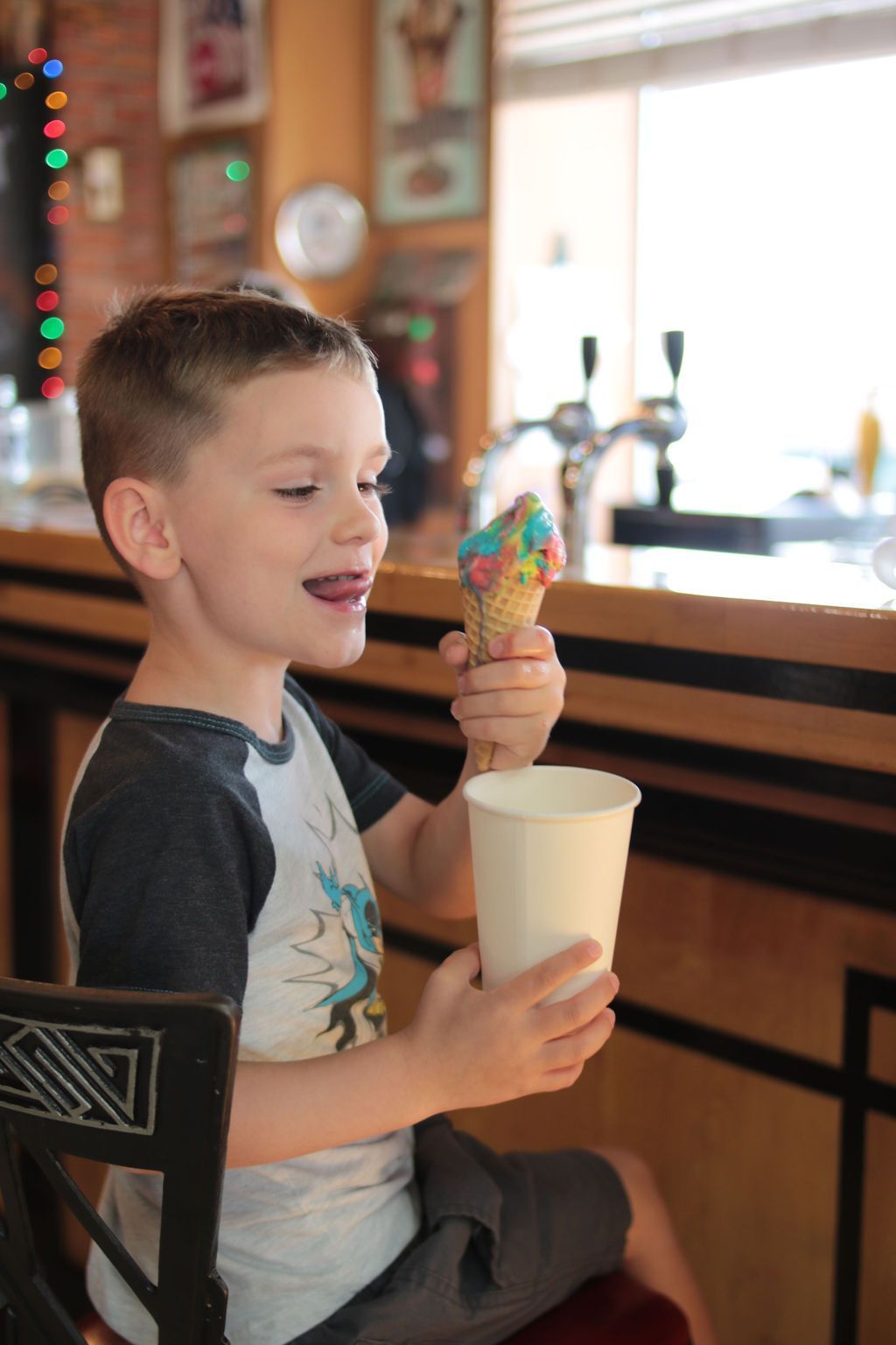 Boy with rainbow ice cream cone, licking his lips, sitting at a counter, holding a cup.