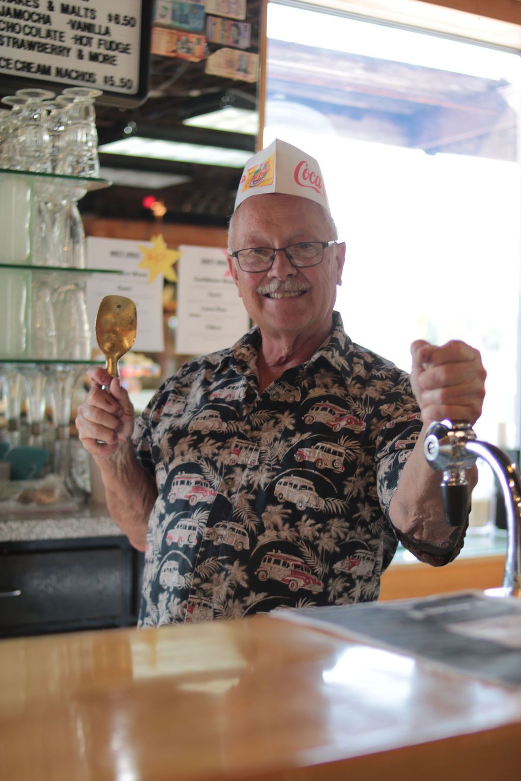 Man in Hawaiian shirt behind counter, holding ice cream scoop, smiling.