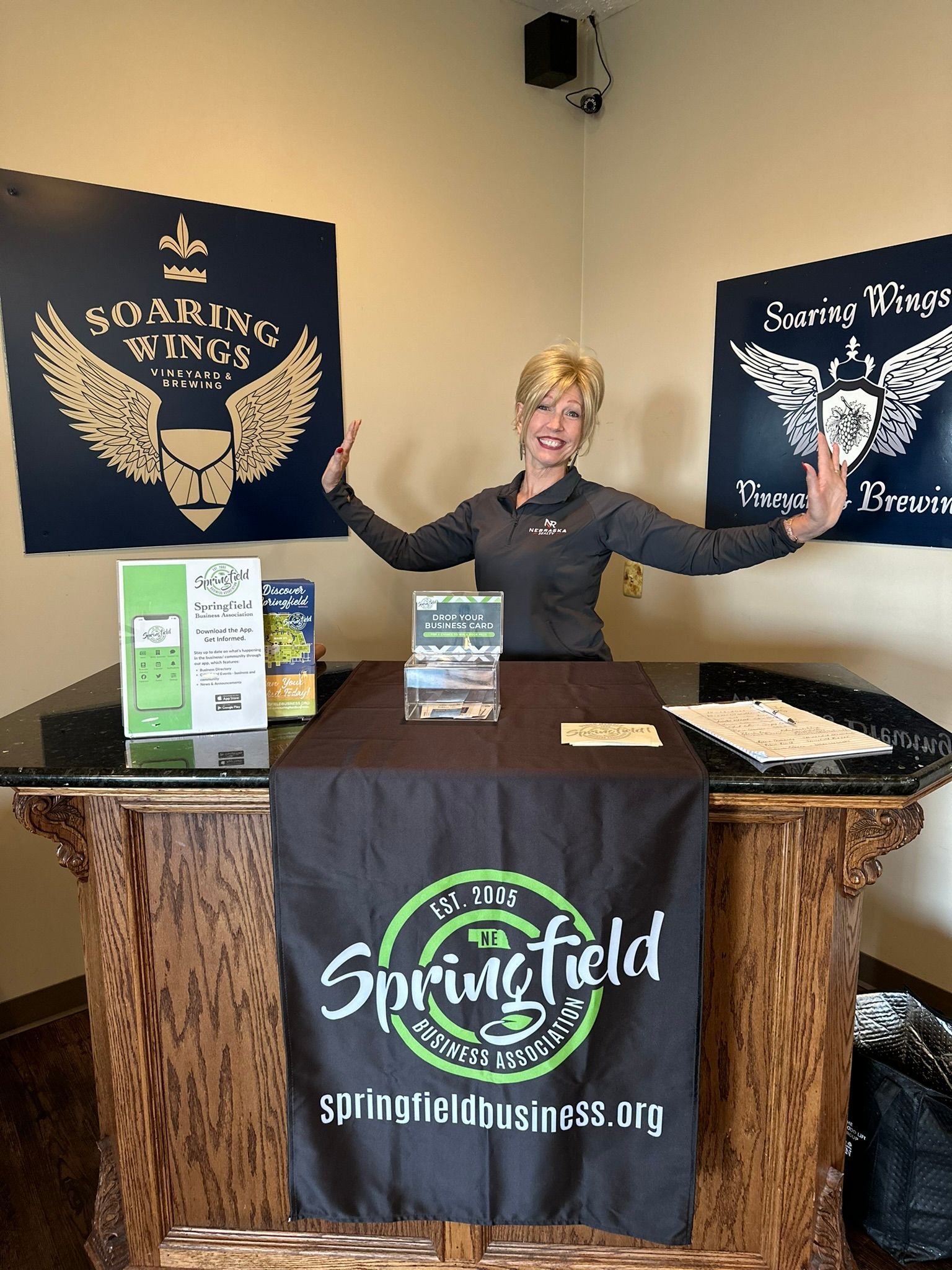 Woman stands at a Springfield, IL business table with arms raised. Soaring Wings sign behind her.