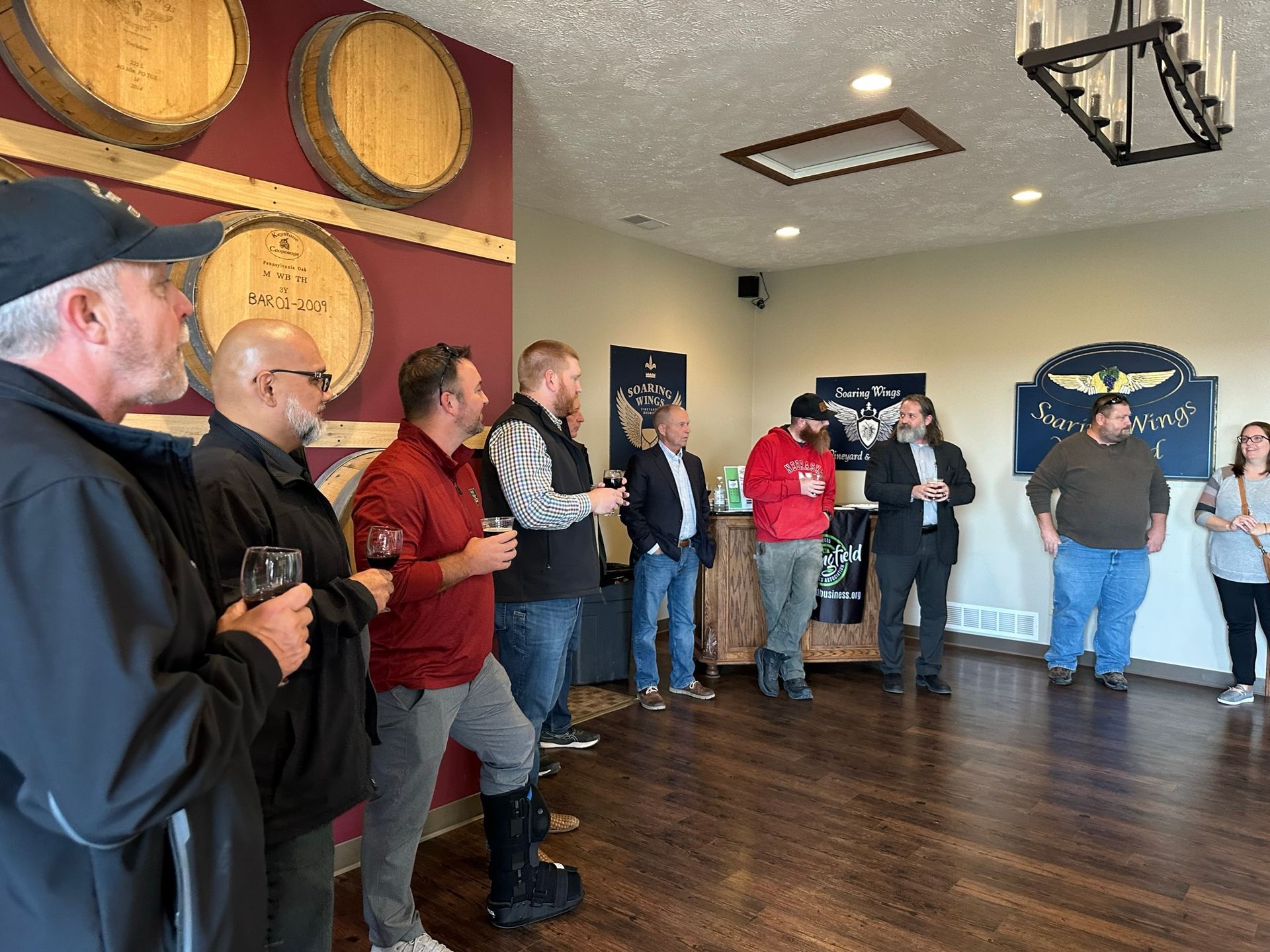 Group of people at an event in a wine cellar, some holding glasses, listening to a speaker. Wine barrels on the wall.