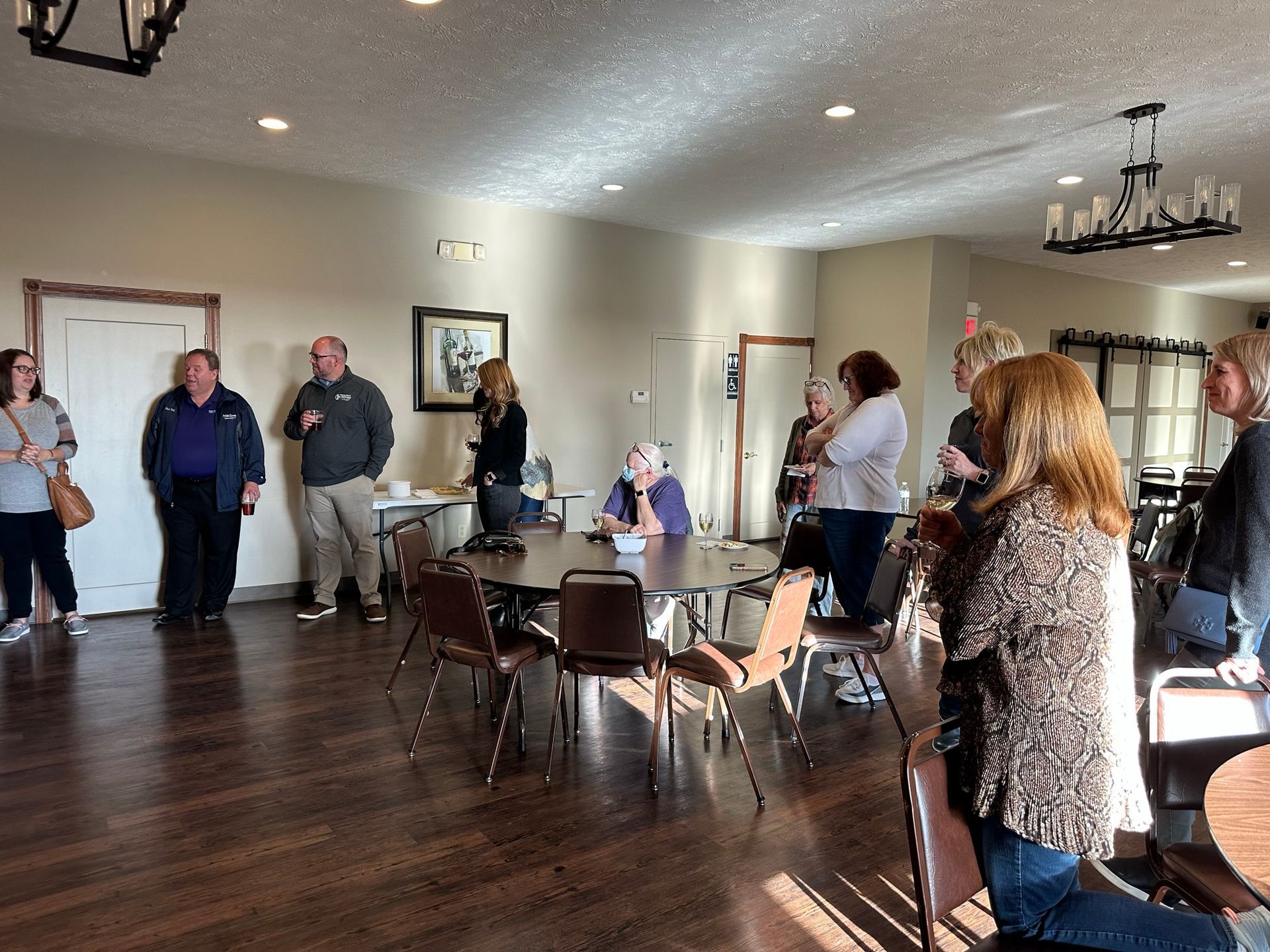 People gathered in a well-lit room with tables, some are standing near doors, and some are interacting at a table.