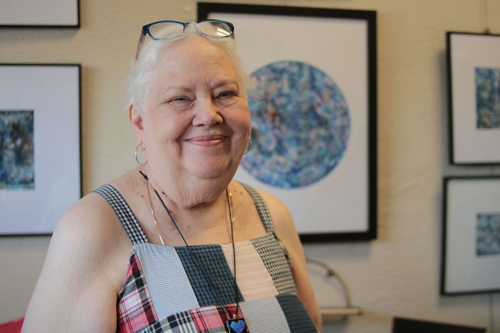 Woman smiles, wearing patchwork top, glasses on head, in art gallery with framed paintings.