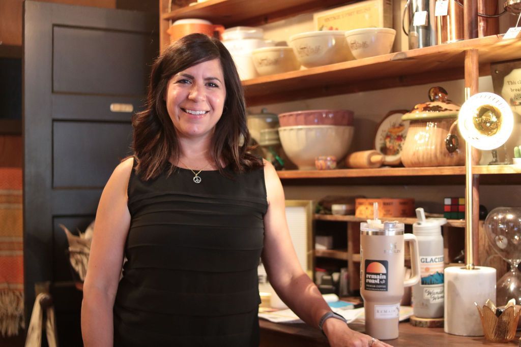 Woman smiling, standing in a shop with shelves of items. She is wearing a black dress.