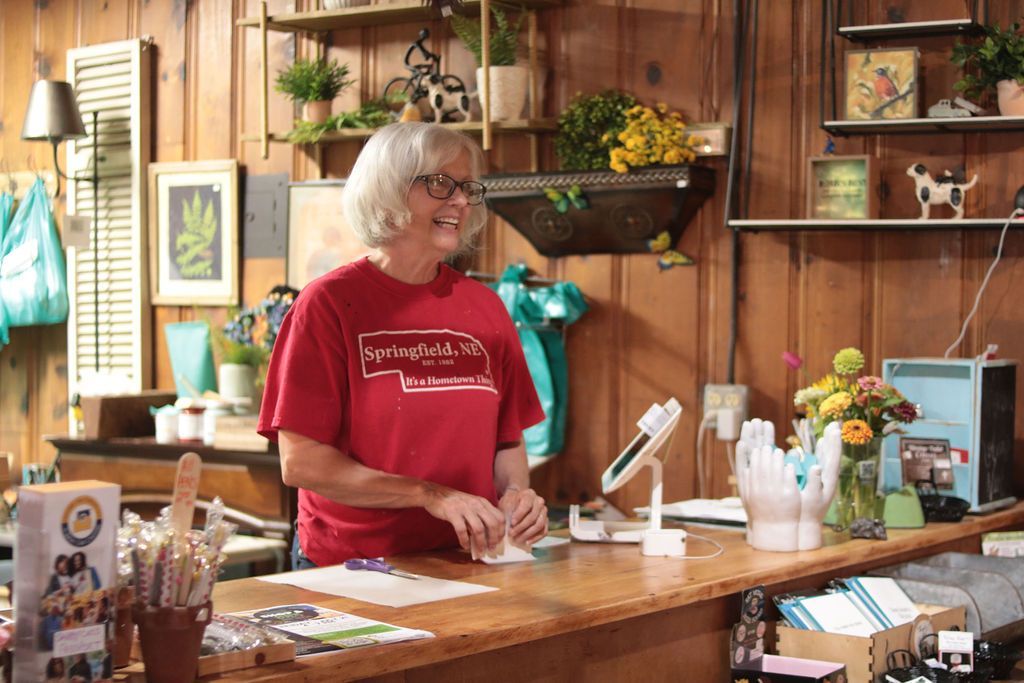Woman behind a counter in a shop, smiling. Wood paneling, plants, and shelves filled with items in the background.