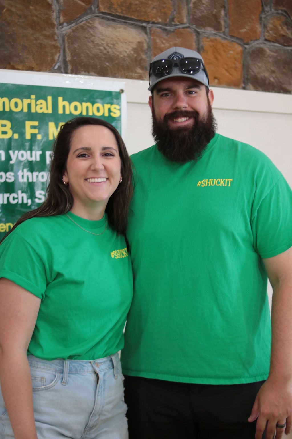 Woman and man in matching green shirts smile, standing near a banner.
