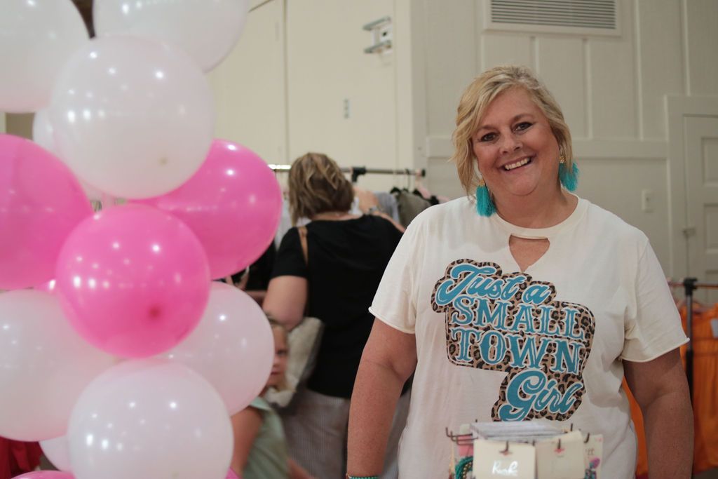 Woman smiles near pink and white balloons; wearing turquoise earrings and a shirt that says 