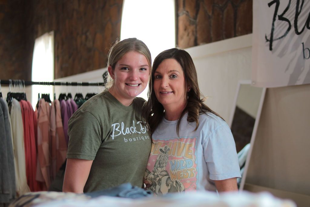 Two women smiling at clothing rack; retail shop setting. One in green shirt, other in blue.