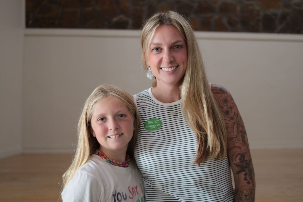 Woman with long blonde hair and a tattoo on her arm, smiles alongside a girl with blonde hair. They stand in a bright room.