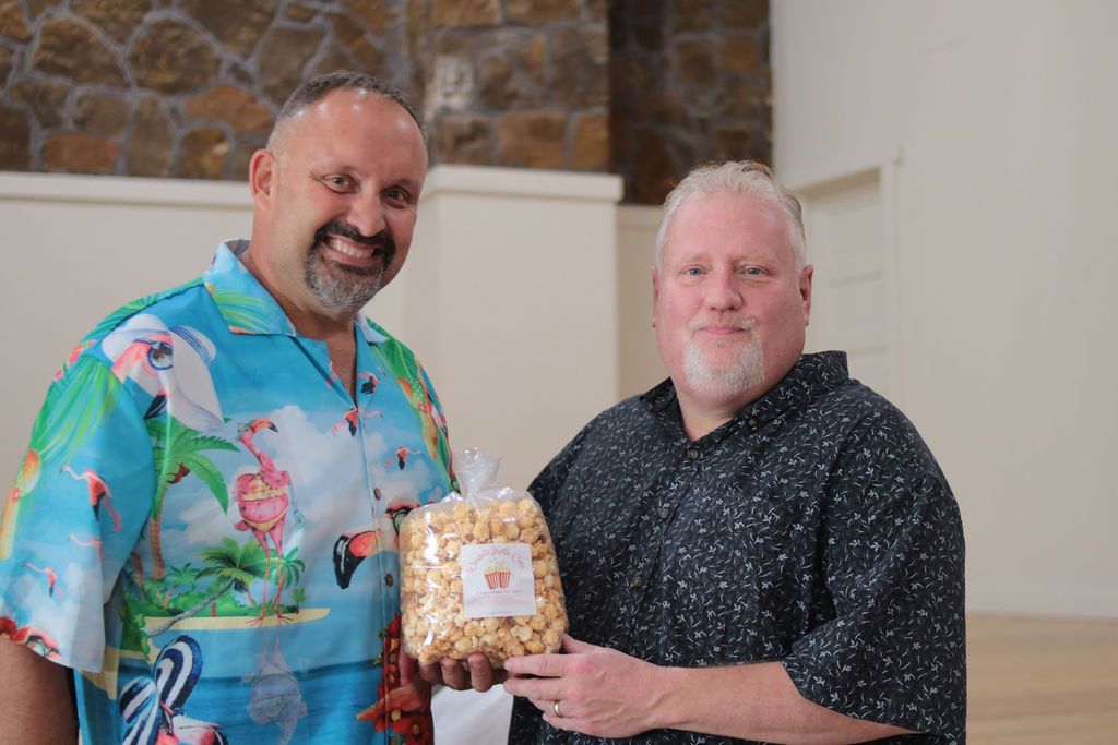 Two men holding a bag of caramel popcorn. One man smiles in a tropical shirt, the other in a patterned shirt. Indoor setting.
