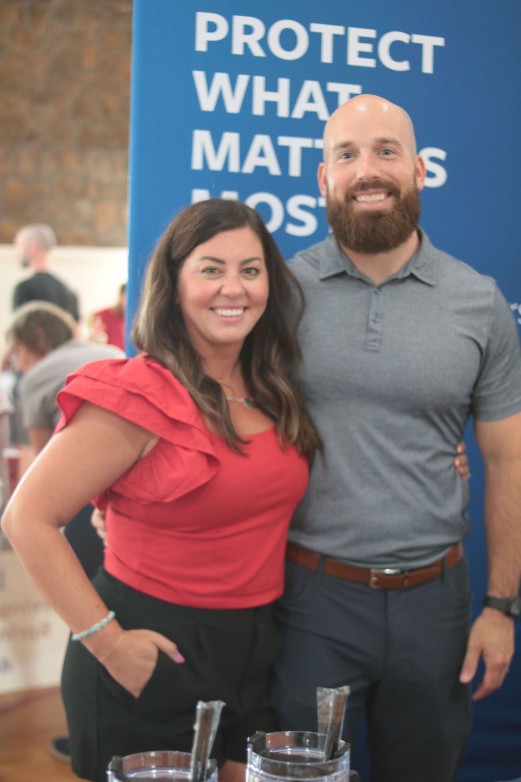 Woman in red top and man with beard pose in front of blue sign.