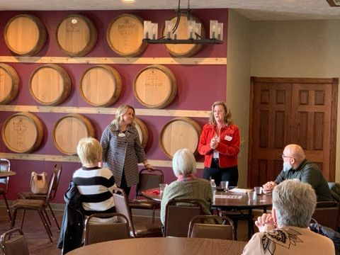 Two women speak to a small group seated at a table. Wine barrels decorate the purple wall.
