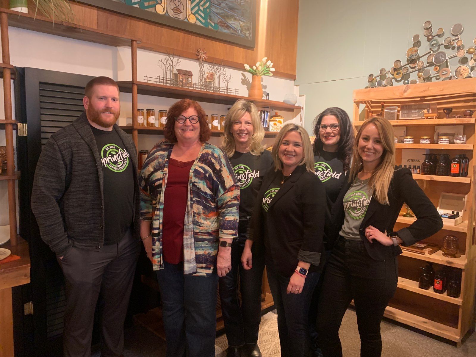 Group of six people smiling in a shop with wooden shelves and art; everyone is wearing a green shirt.