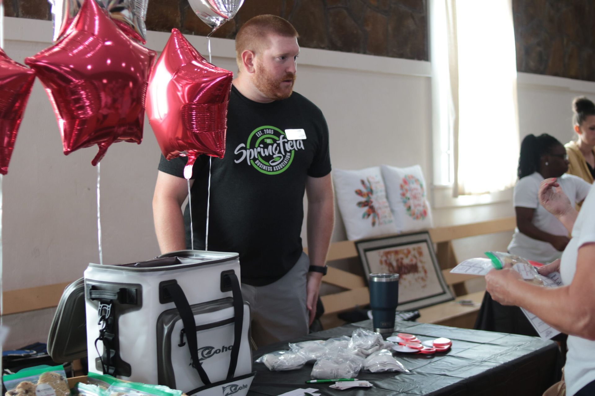 Man at a table with balloons and a cooler. Other people are near. Setting is indoors.