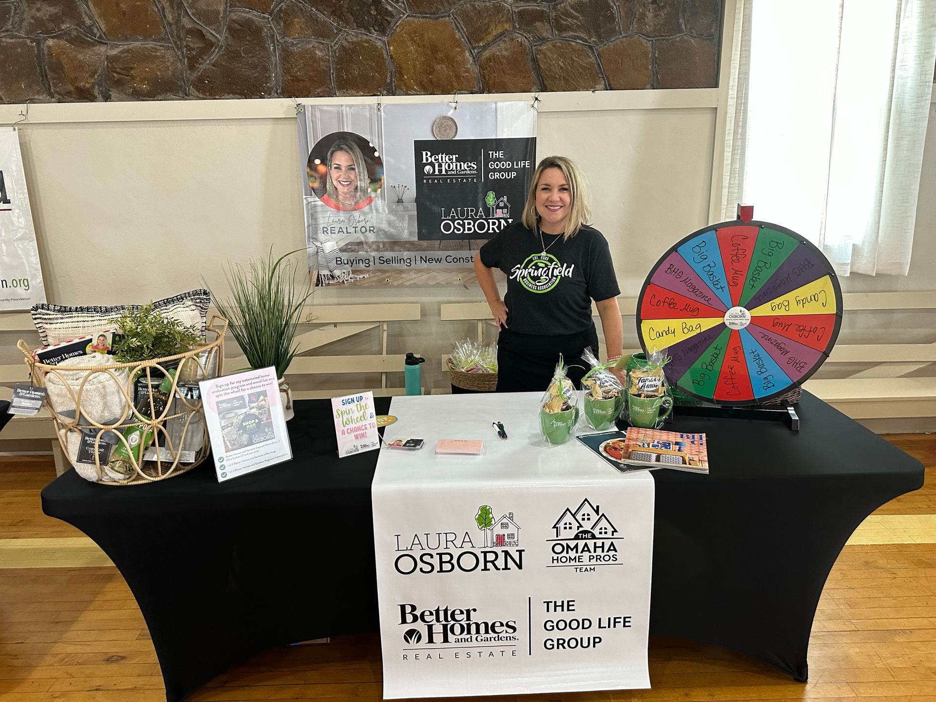 Woman at a Better Homes & Gardens booth with a prize wheel, brochures, and greenery.