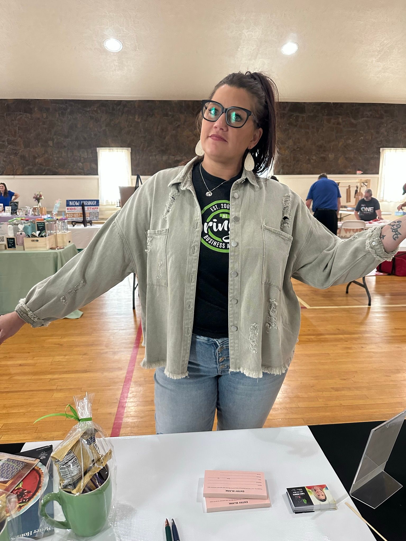 Woman in oversized jacket, arms out, at a vendor table with items. Interior shot with tables, windows, and spectators.