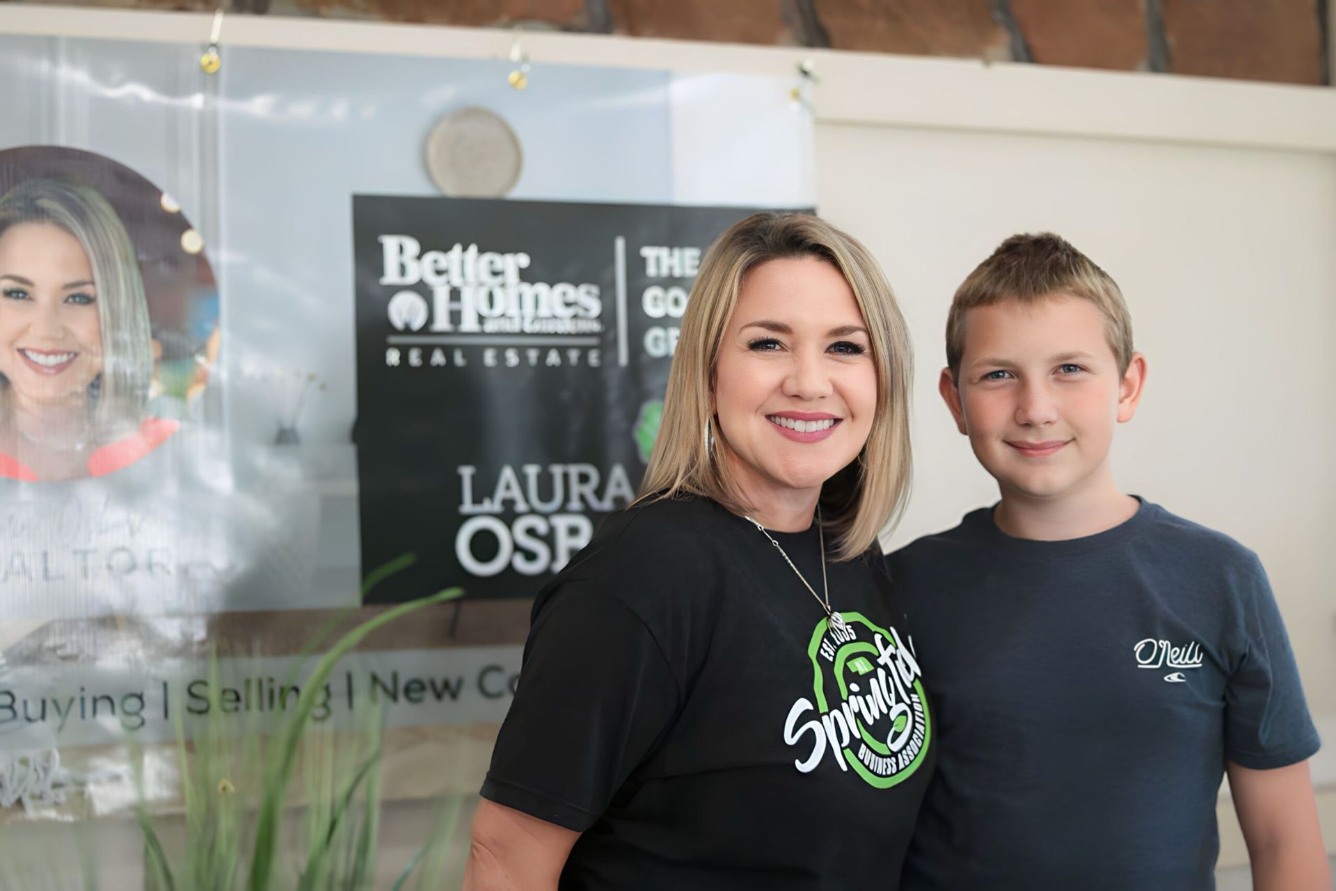 Woman and boy stand in front of a real estate sign. The woman smiles, boy has a neutral expression.