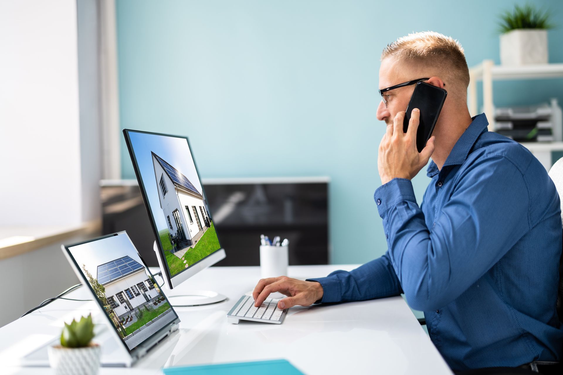 Man in blue shirt on phone at desk with computer monitors in a bright office