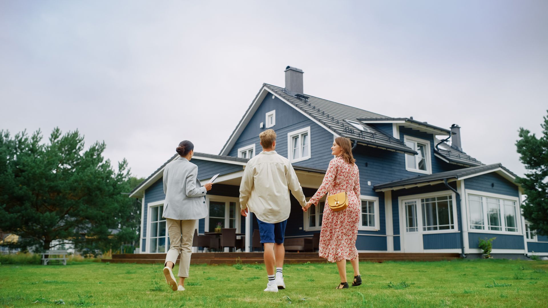 Three people walking hand in hand toward a blue house with a lawn and cloudy sky
