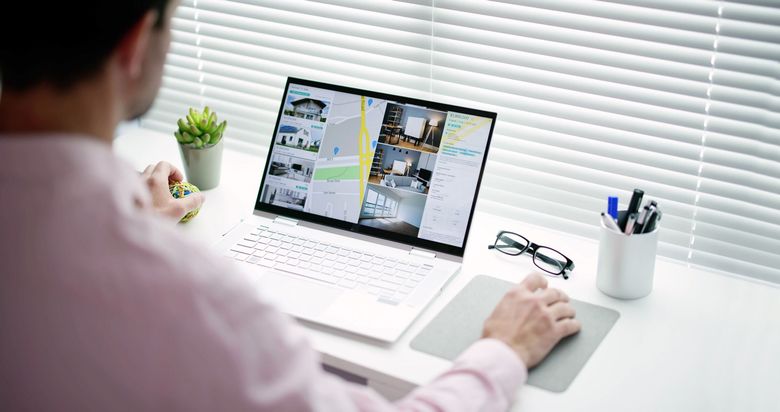 Person working on a laptop at a white desk, reviewing a website, with glasses, plant, and pen holder nearby