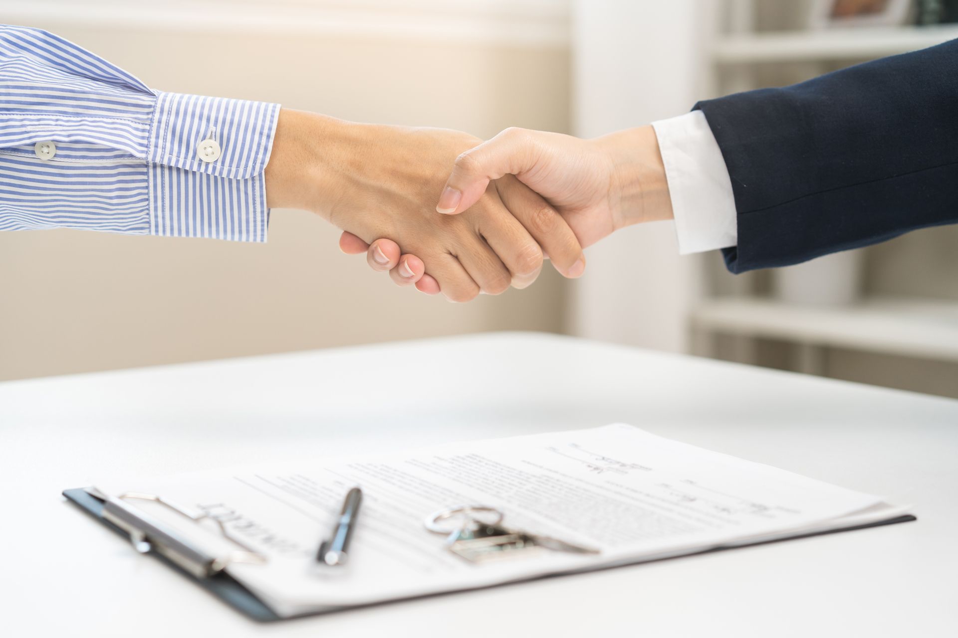 Two people shake hands over a desk with signed paperwork and pens.