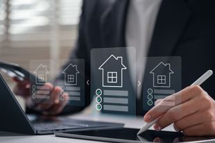 Hands using a stylus and tablet with holographic house icons over a desk