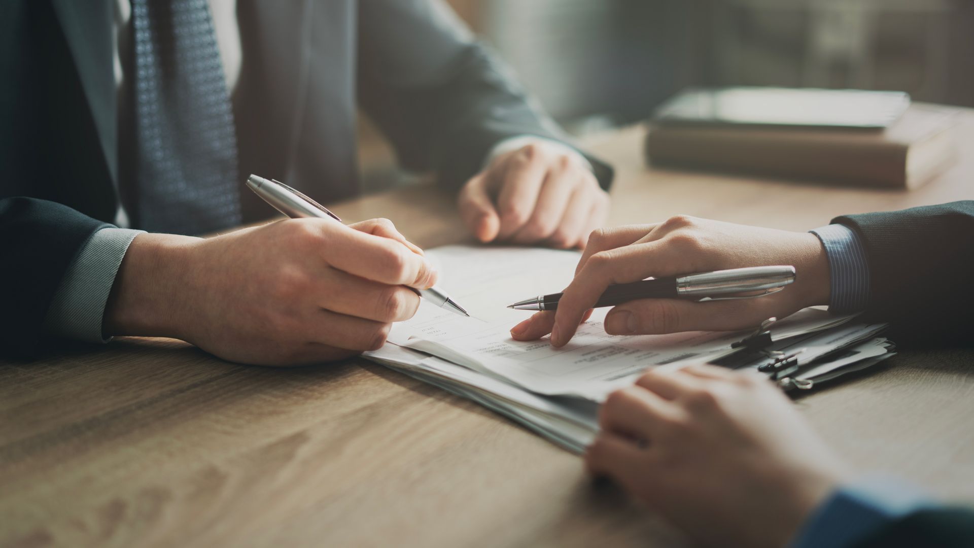 Hands signing a document at a wooden table with pens and papers, in a meeting setting