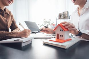 Two people reviewing a house model and documents at a desk with a laptop