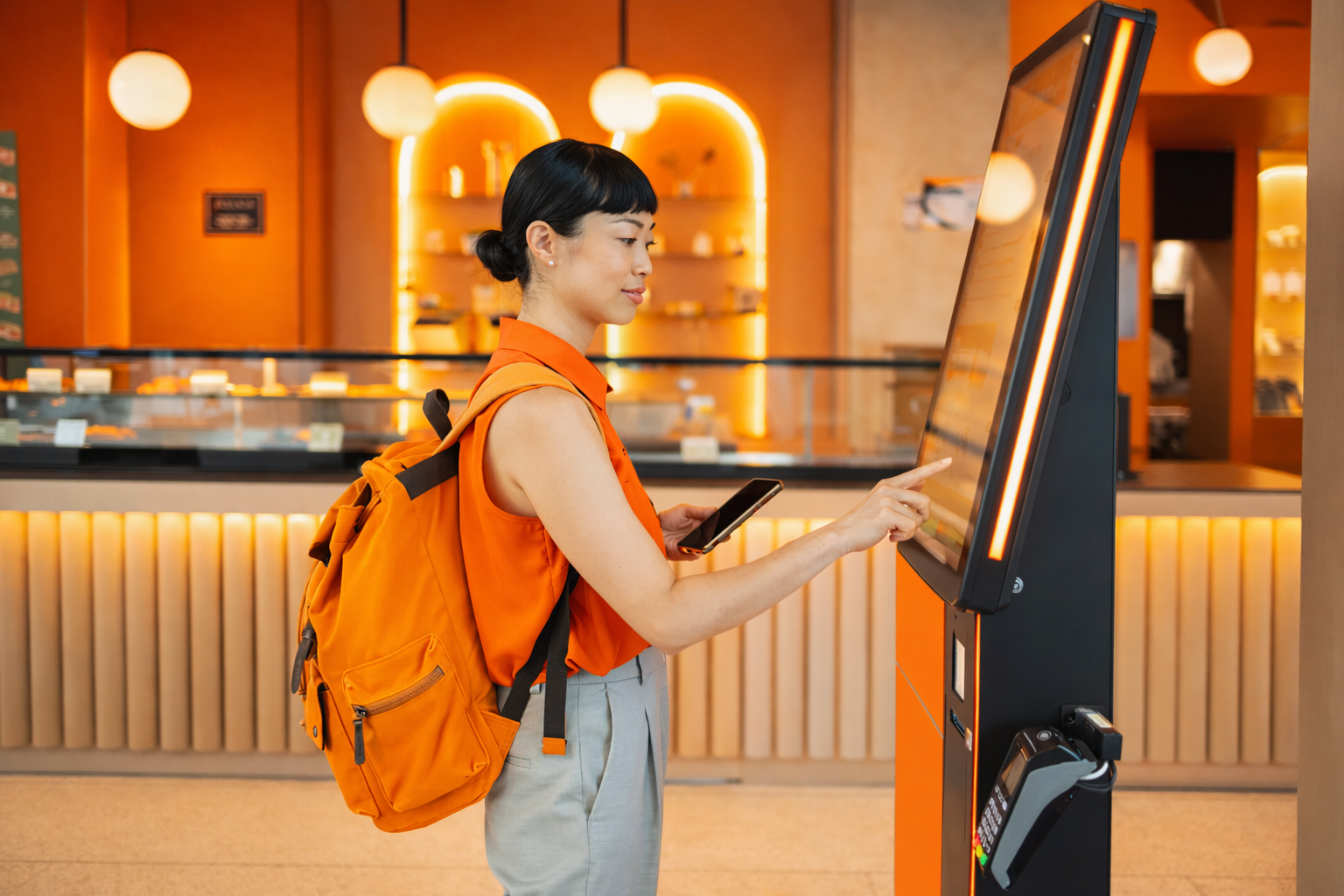 Person with orange backpack uses a kiosk in a warmly lit lobby